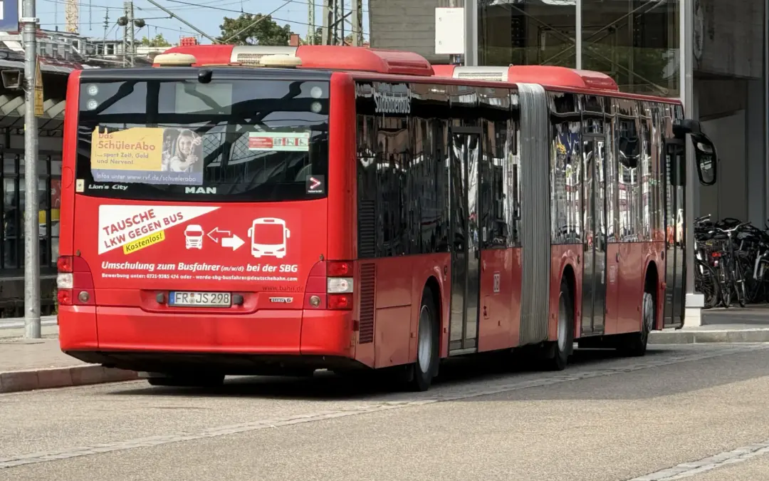 Rear and side view of a bright red city bus parked along a curb in an urban area, with reflective windows and overhead wires nearby.