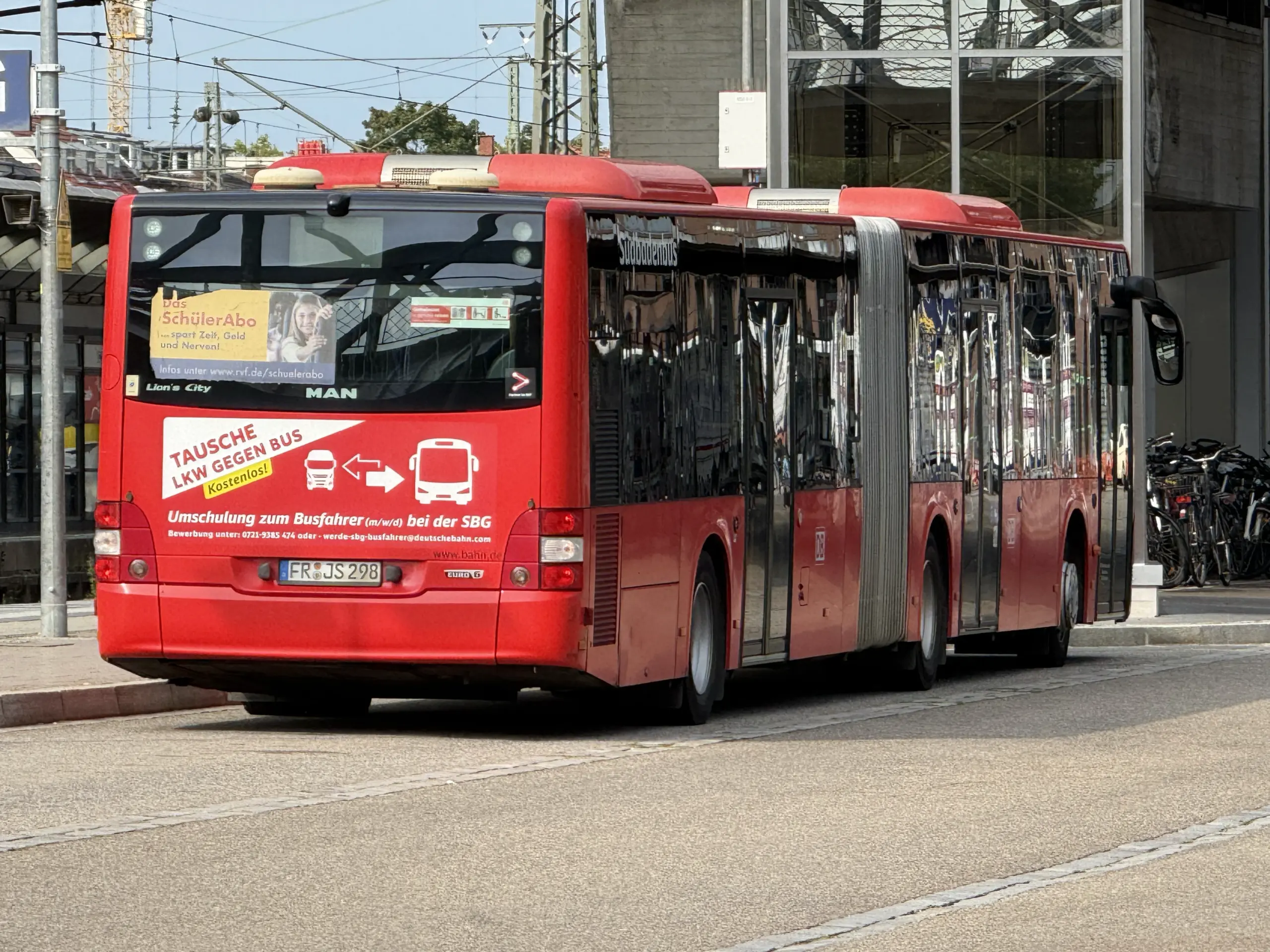 Rear and side view of a bright red city bus parked along a curb in an urban area, with reflective windows and overhead wires nearby.